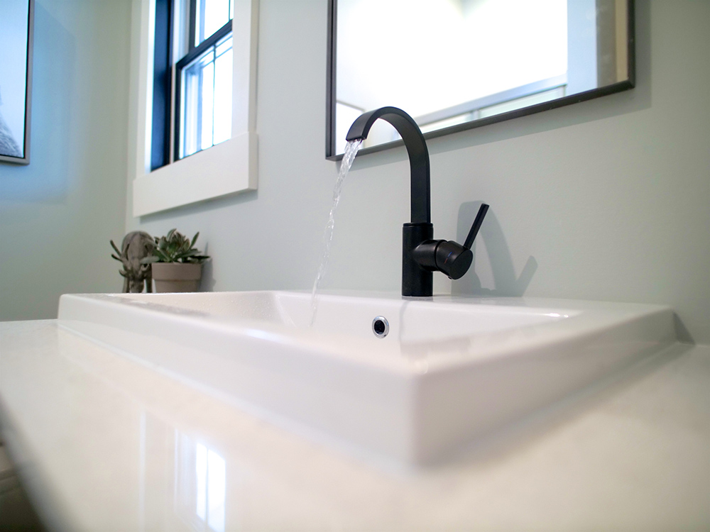 A bathroom with a white sink with black faucet, a mirror, window and plant.