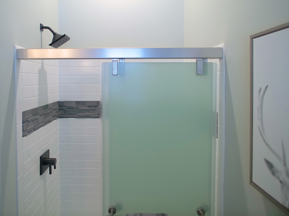 Shower with a glass door, shower head, grey and white tile, and wall art.