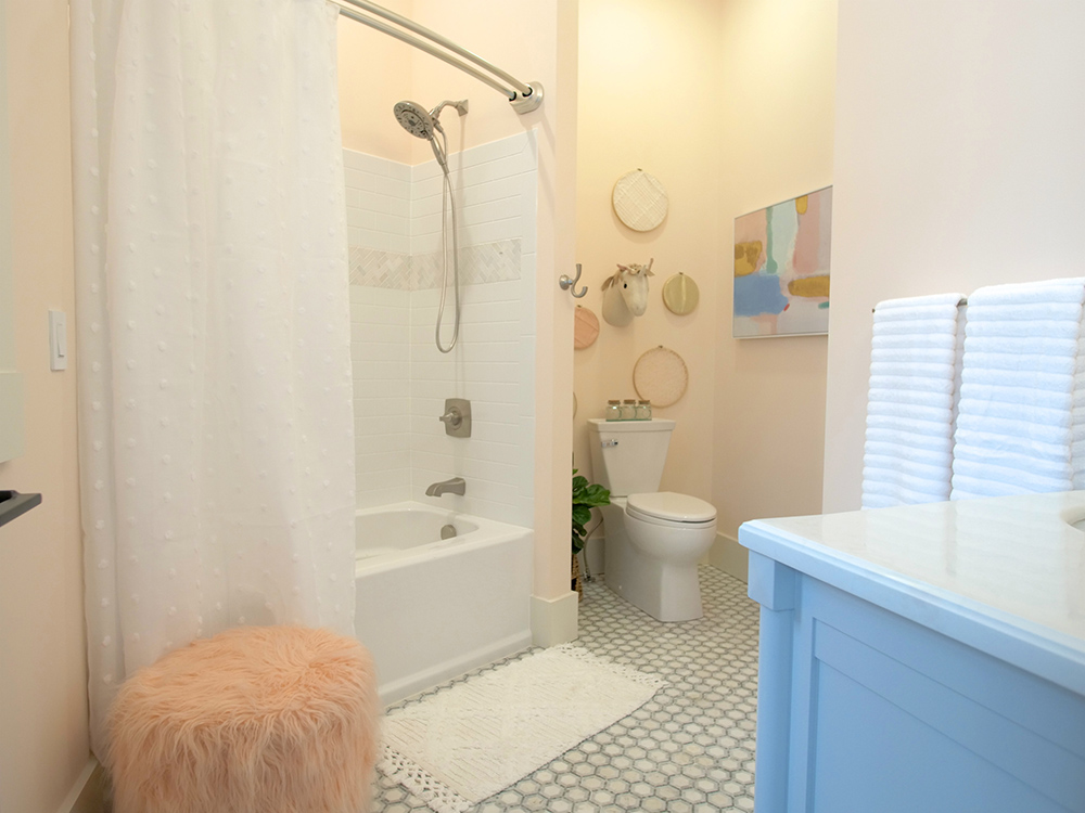 A bathroom with a white shower curtain, grey and white tile floor, white toilet, light grey blue vanity, wall art and a pink stool.