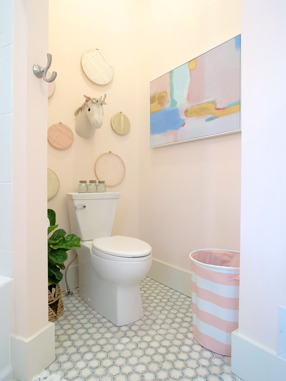 A white toilet, white and grey tile, peach colored walls, wall art and a pink and white laundry basket.