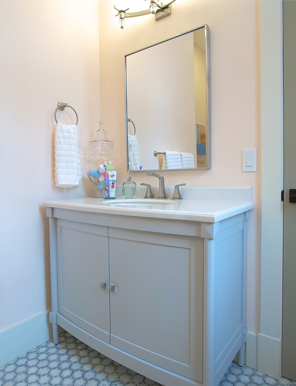 A grey blue vanity, mirror, silver appliances, white hand towel, and white and grey tiled floor.