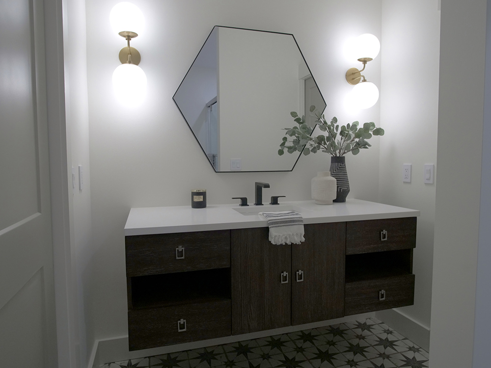 A bathroom with white walls, a wooden and white vanity, a hexagon shaped mirror, two sconces, white linens and a plant.