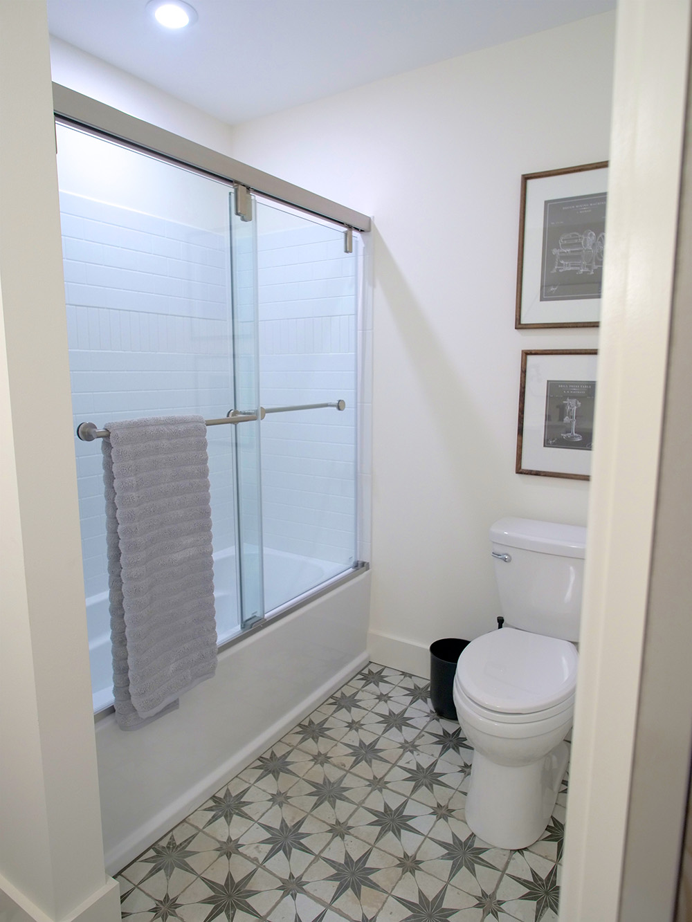 A bathroom with a shower and glass door, grey towel, white toilet, framed wall art and grey and white tile flooring