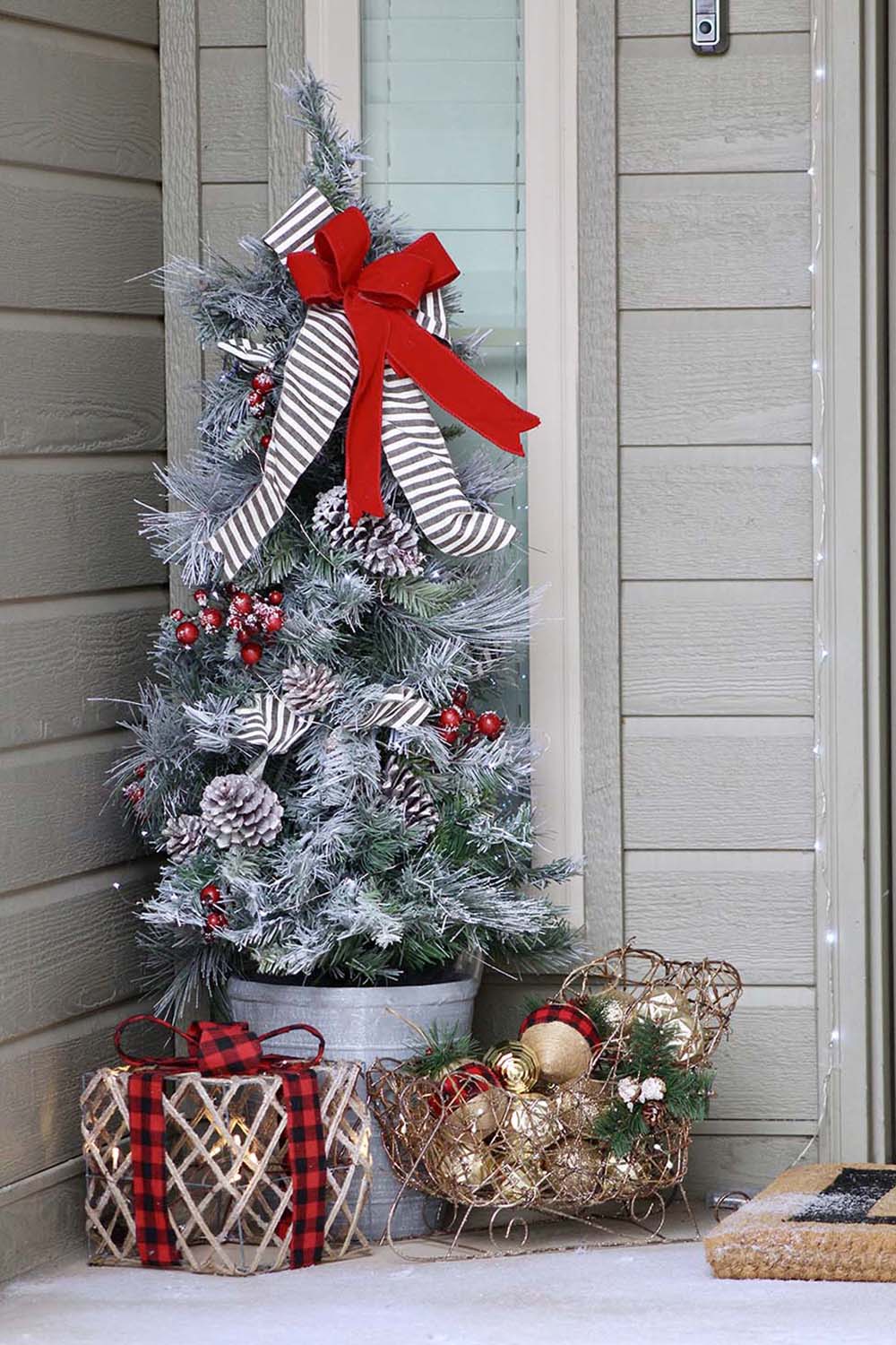 The corner of a front porch decorated with a porch Christmas tree, lighted holiday gift box, and ornament filled sleigh.