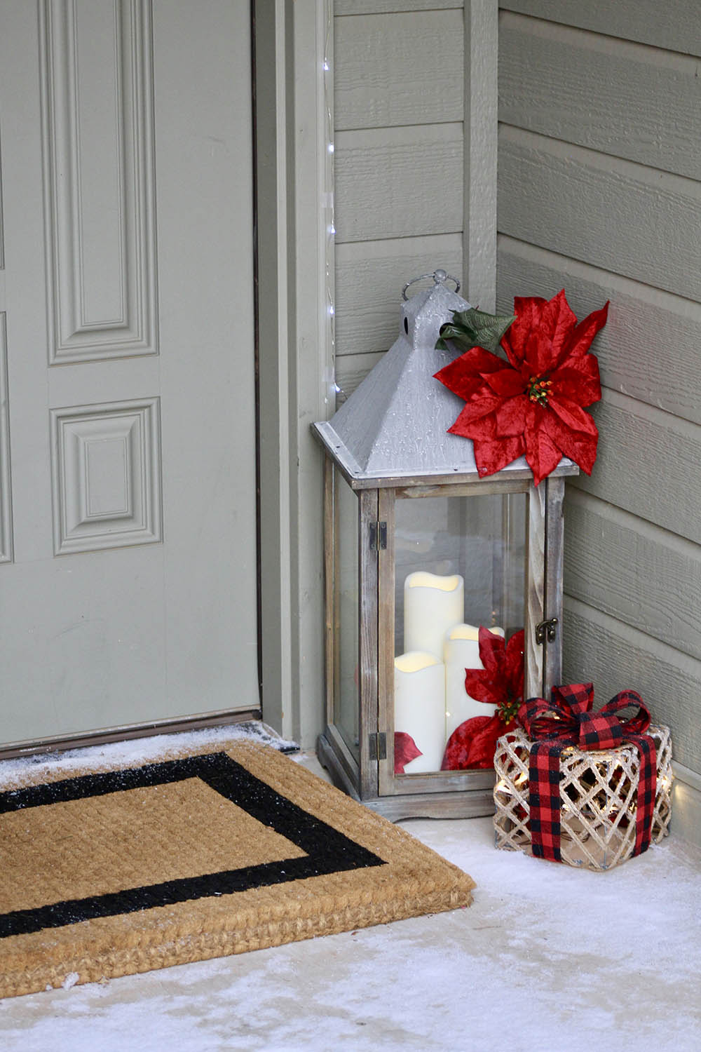 A large metal lantern filled with LED candles and a lighted holiday gift box decorates the corner of a porch. 