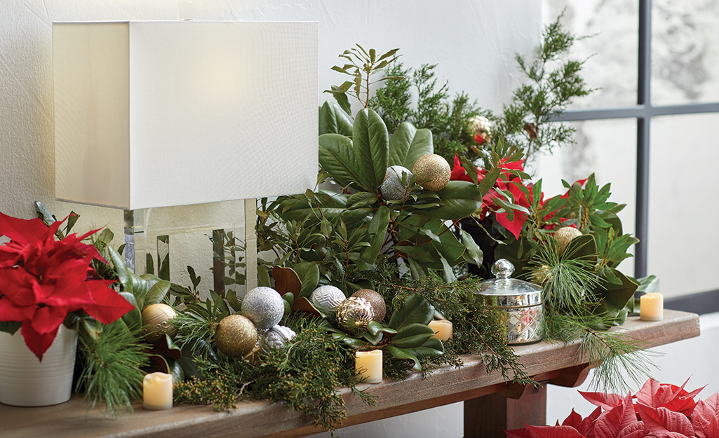 Fresh greenery embellished with metallic Christmas ornaments and candles on a side table.