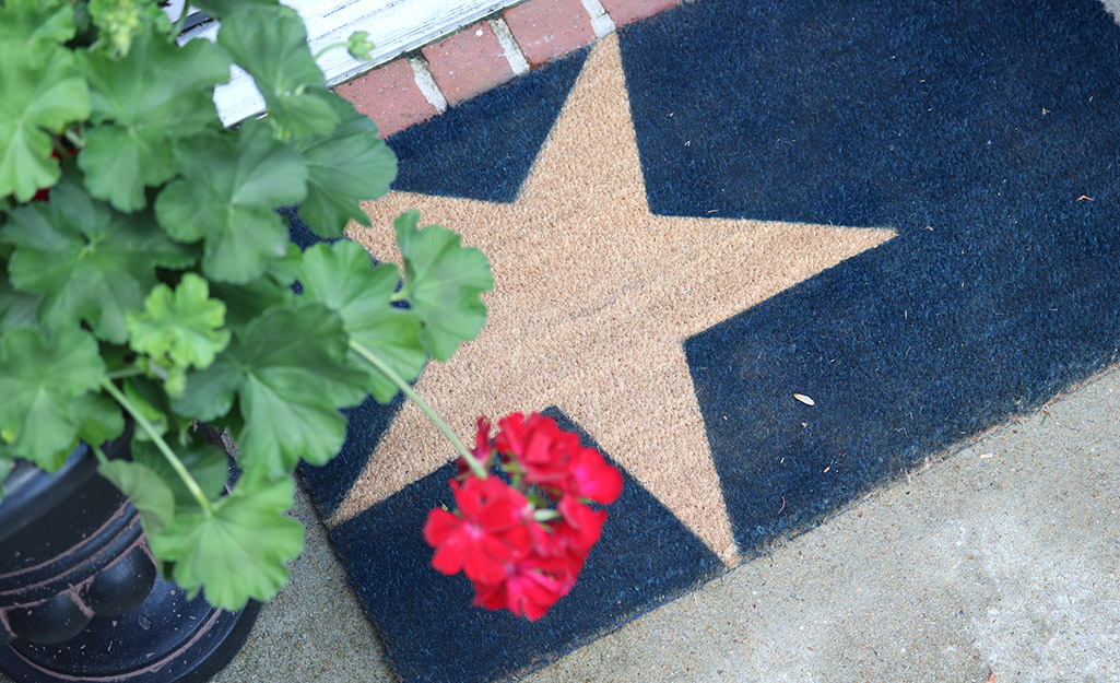 A door mat with painted star lays next to a geranium outside a house.