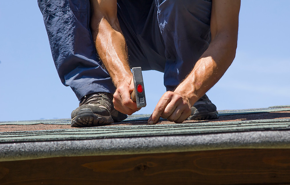 A person standing on a roof holds a hammer while checking roof shingles.