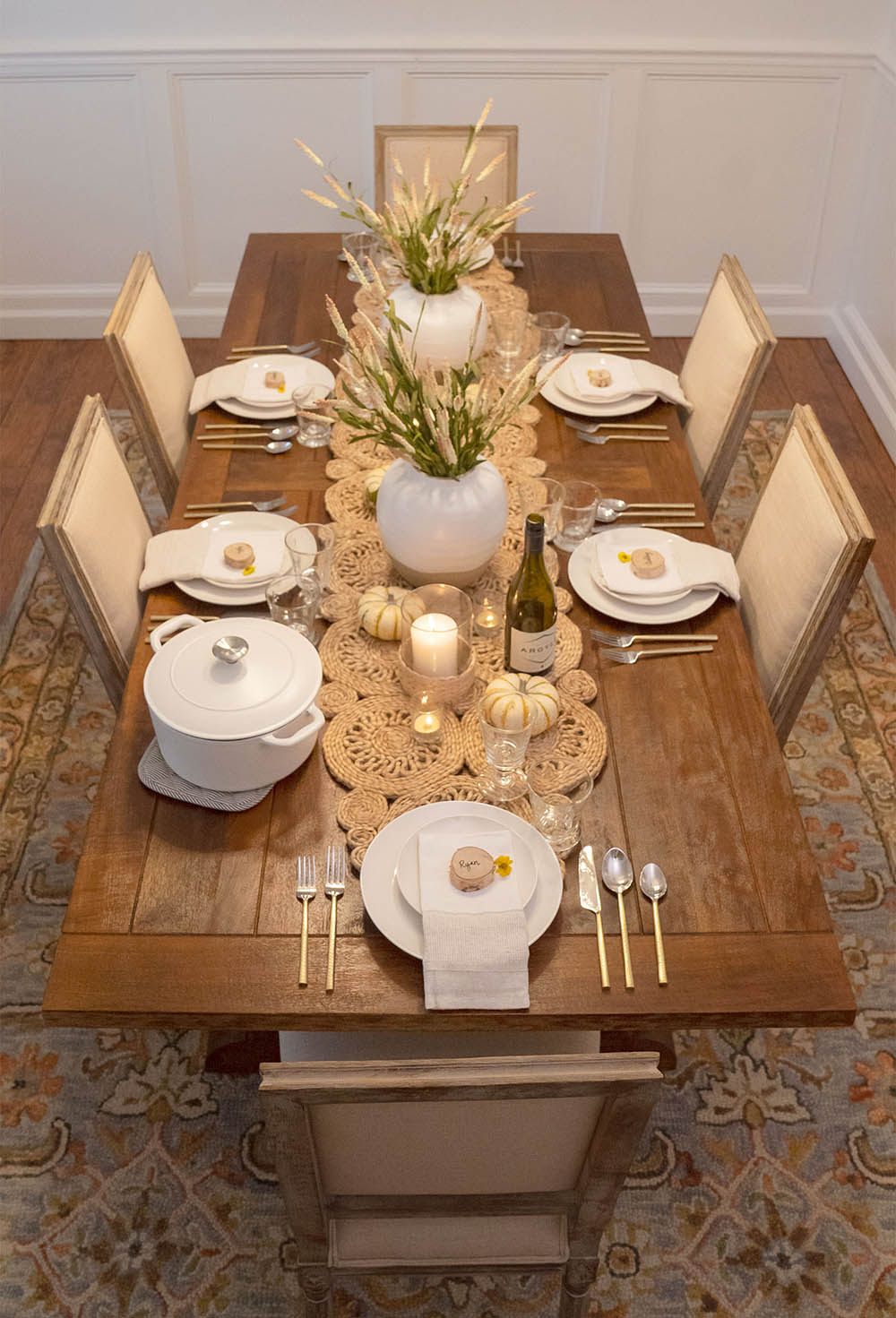 A wooden dining room table decorated with white dishes, silver flatware, candles, and miniature pumpkins for Friendsgiving.