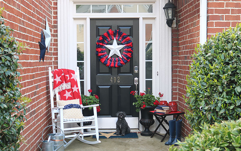 A 4th of July wreath hangs above a door sitting by the front door of a house with an entranceway decorated for Independence Day. 