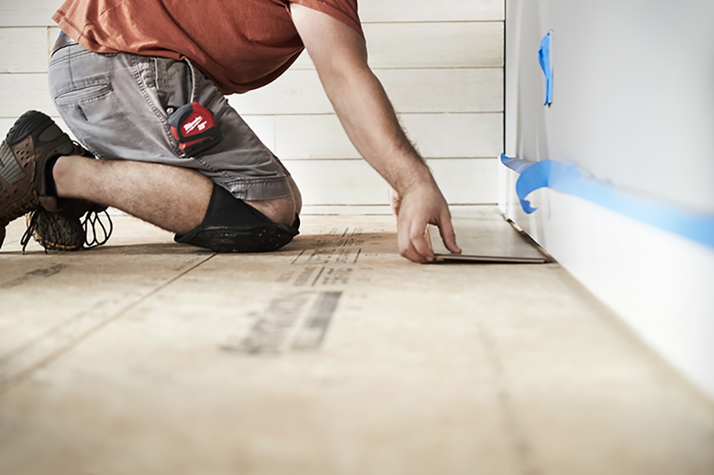 A person kneeling on a subfloor lays a laminate flooring plank against a baseboard.