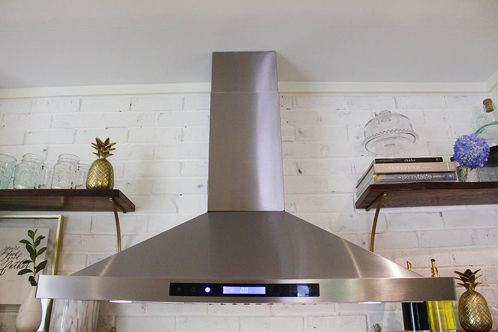 A limewashed wall with a stainless steel hood vent surrounded by open shelving.