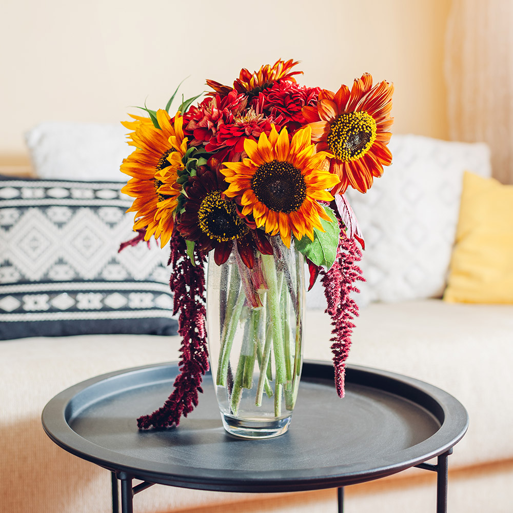 A vase of orange and yellow sunflowers accented by red and purple flowers stands on a small round table in front of a sofa.