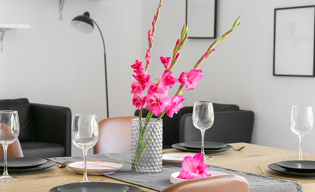 Three stems of pink gladiolus stand in a patterned glass vase at the center of a table set with wine glasses and black plates.