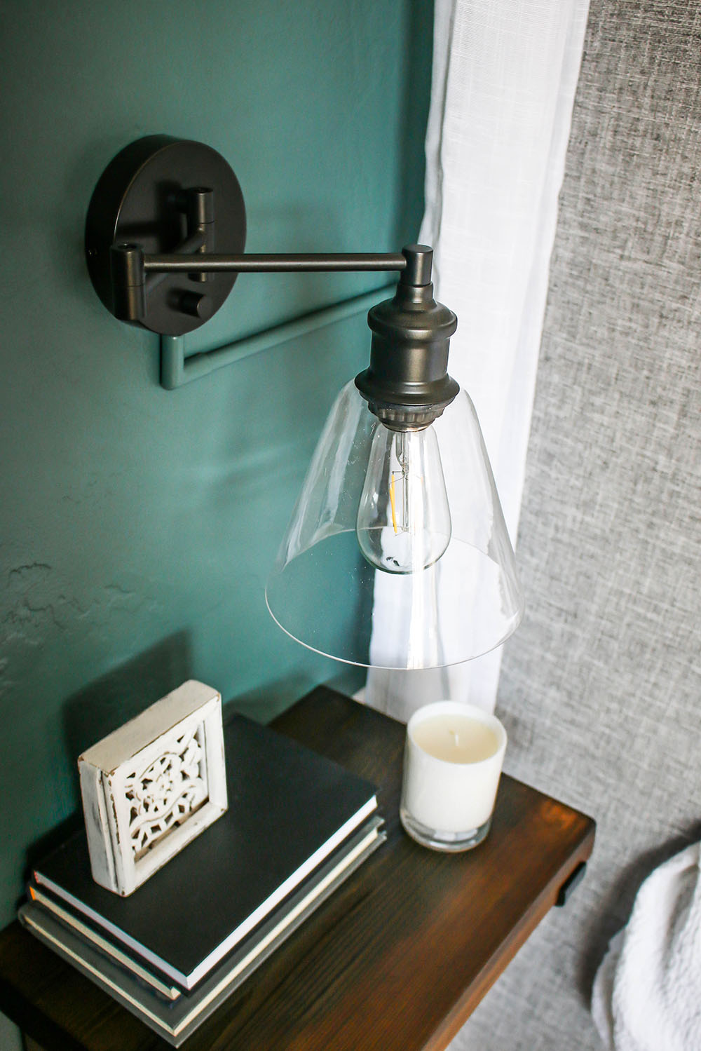 A black sconce on a dark green wall and a wooden table with books and a candle. 