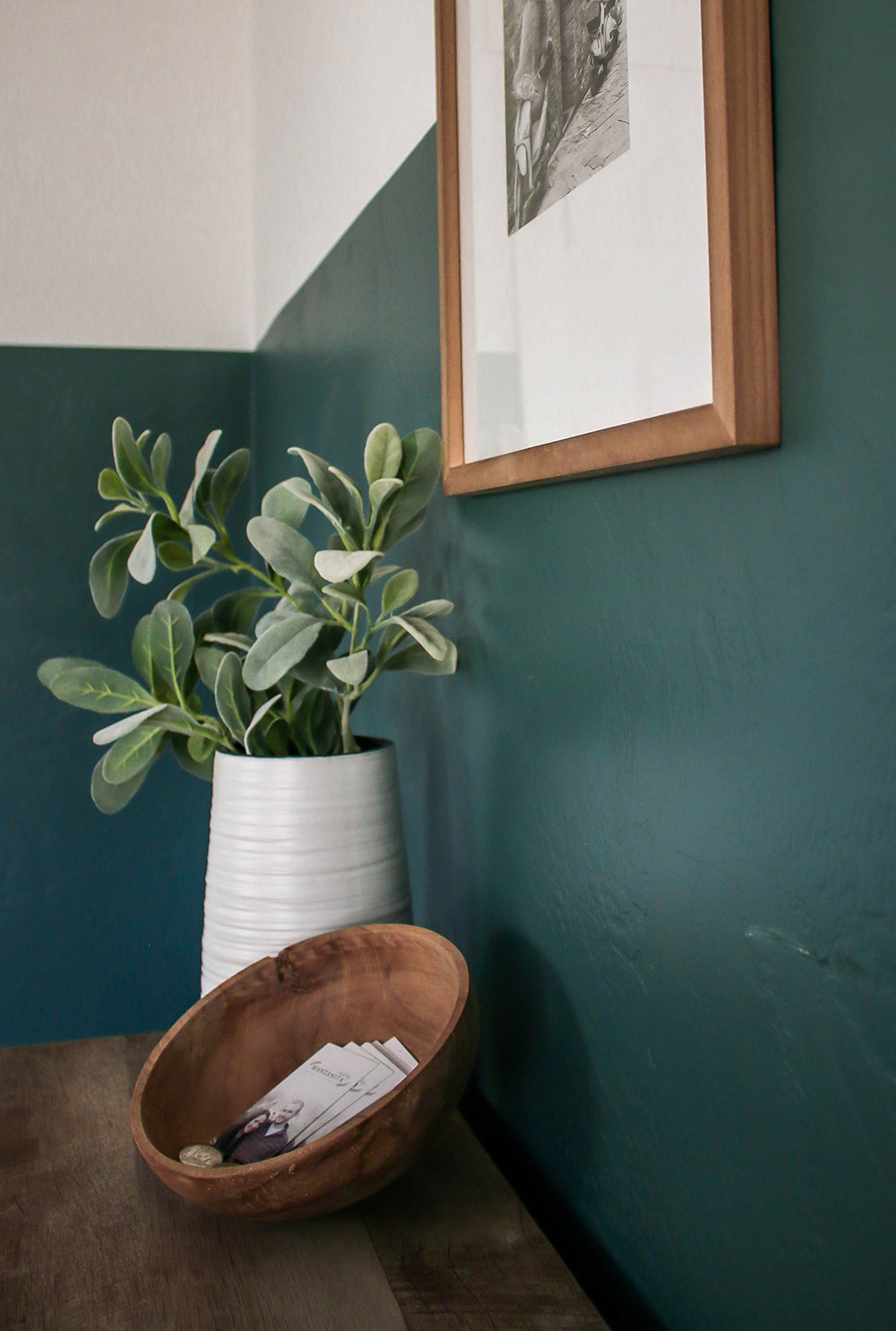A dark green and white wall with a framed piece of art work and a wood table with a white vase and wooden bowl.