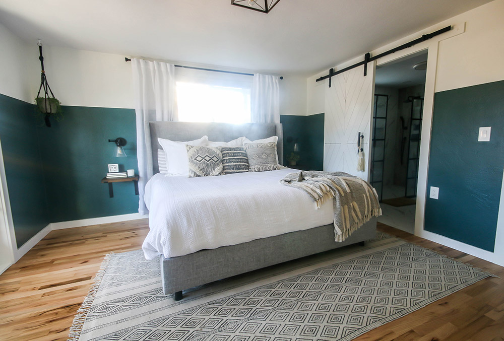 A bedroom with wood flooring, a grey and white rug, a bed with a white comforter and throw pillows, one hanging light fixture, a window with white drapes, a white sliding barn closet door, and dark green and white walls.