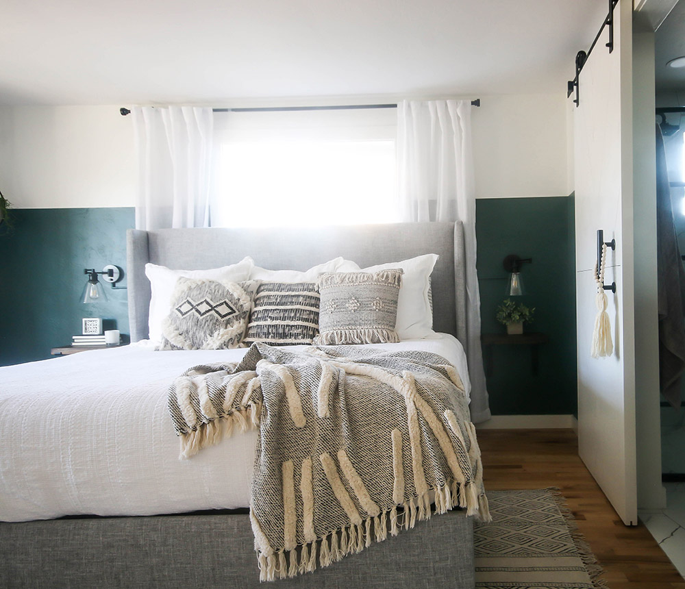 A bedroom with wood flooring, a grey and white rug, a bed with a white comforter, throw pillows and a blanket, two sconces, a window with white drapes, a white sliding barn closet door, and dark green and white walls.