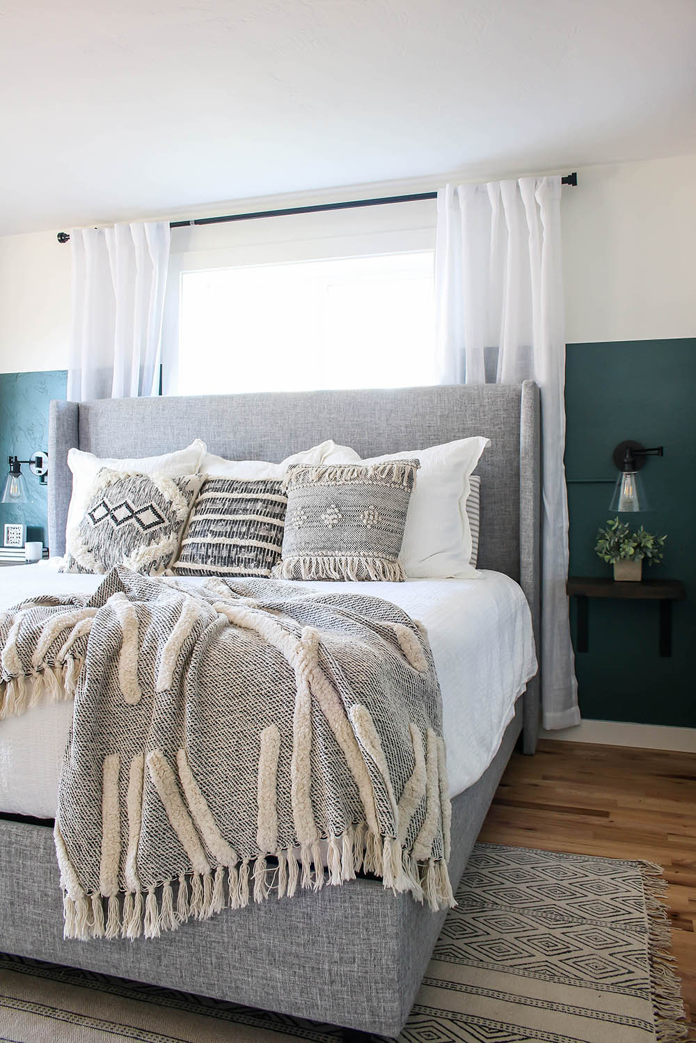 A bedroom with wood flooring, a grey and white rug, a bed with a white comforter, throw pillows and a blanket, two sconces, a window with white drapes, and dark green and white walls.