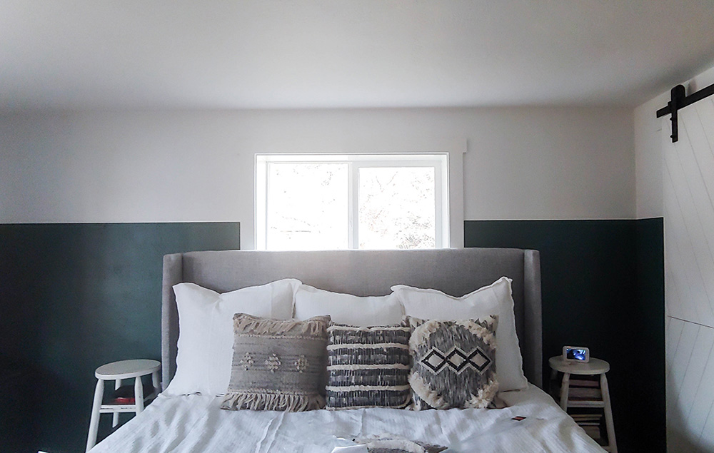 A bedroom with dark green and white walls, a bed with a grey headboard, throw pillows and a white comforter, and two side tables.