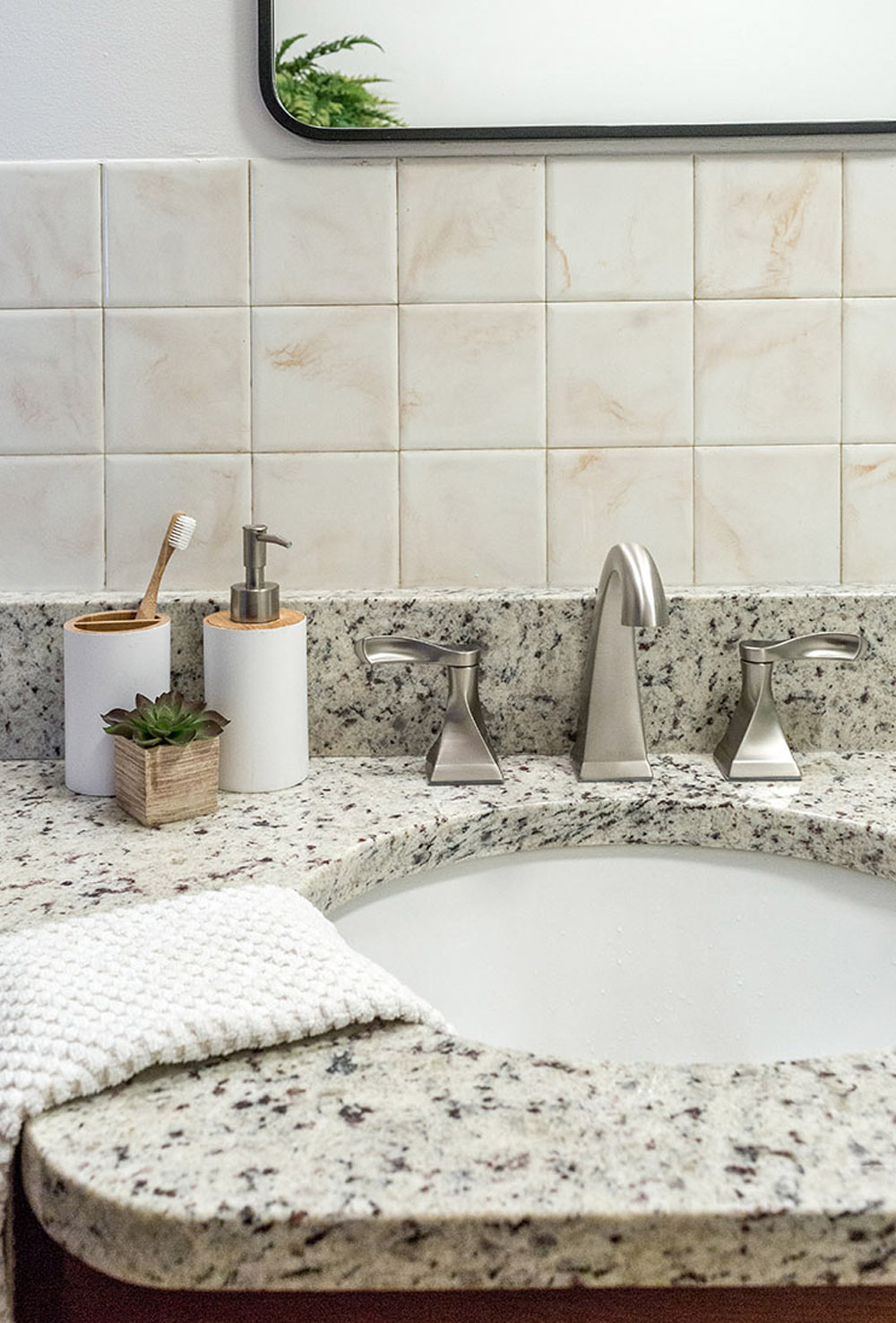 Marble bathroom countertop with a white hand towel, silver appliances and white soap dispenser, cream wall tile and a mirror.