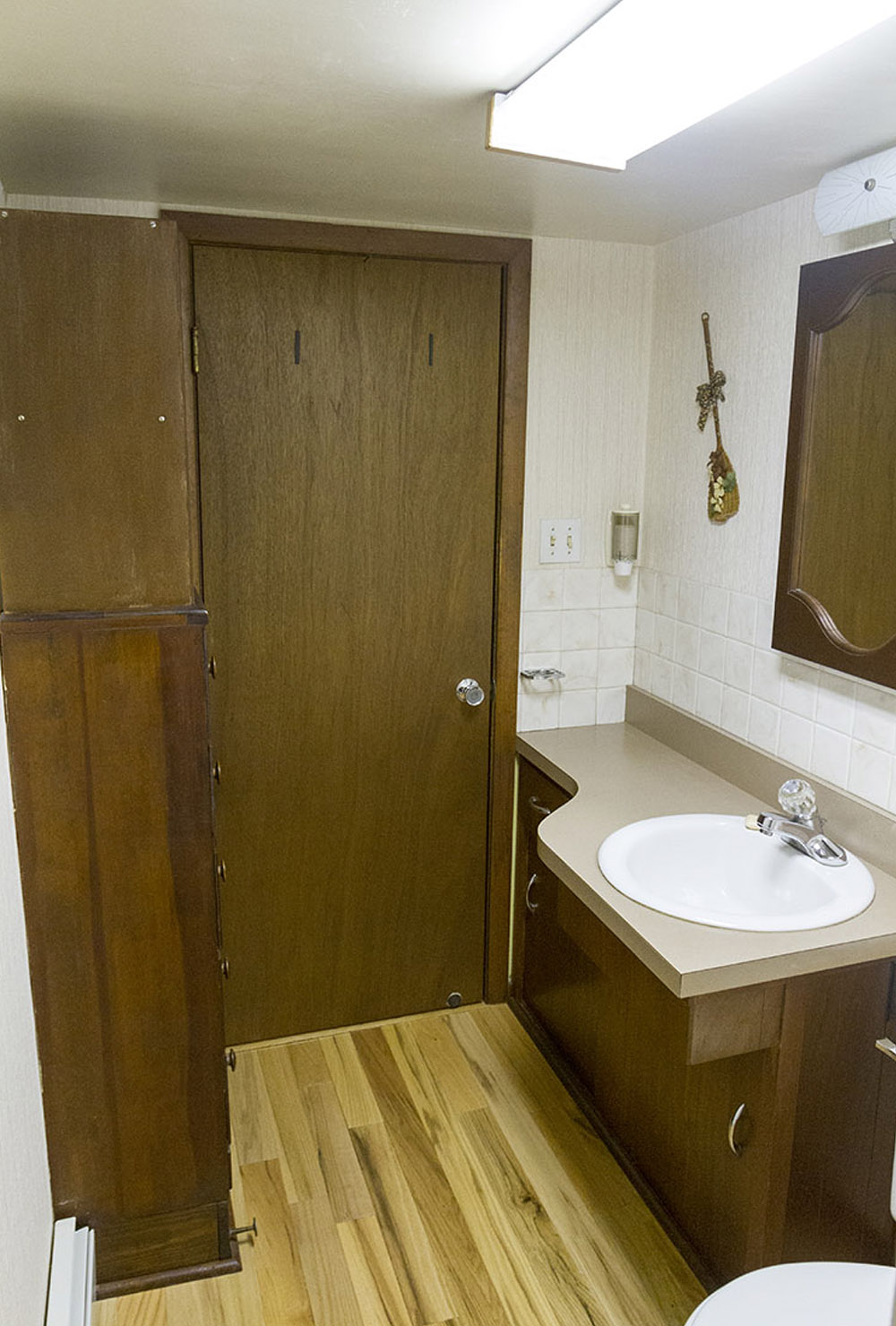 A bathroom with a brown wooden door, wooden cabinet, brown vanity, wood flooring and a beige, tiled wall with a mirror.