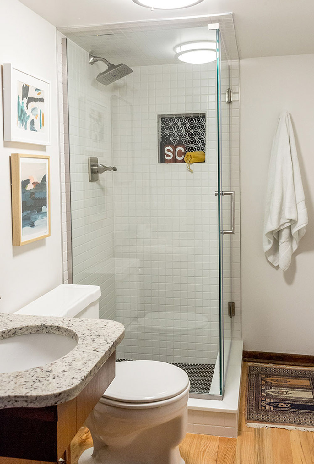 A bathroom with white walls, a glass shower, white toilet, wooden vanity, mirror, and gold bathroom light fixture.