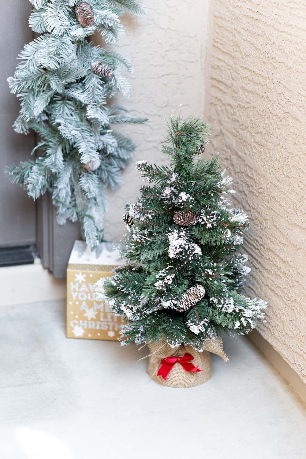 A front porch is styled with garland, a present and a small tree shot in corner