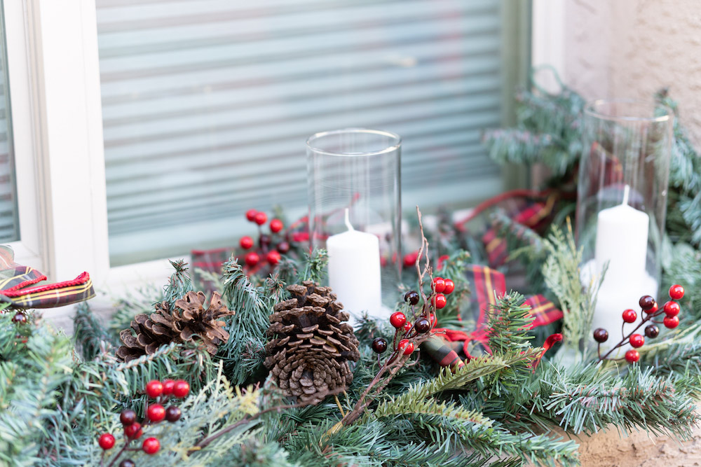 A window sill is styled with garland, pinecones, and candles
