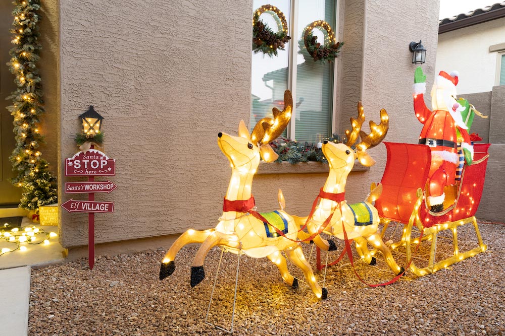 A front porch is styled with a holiday sign, garland, wreaths, a small tree, lights and an illuminated LED Santa display with reindeer  