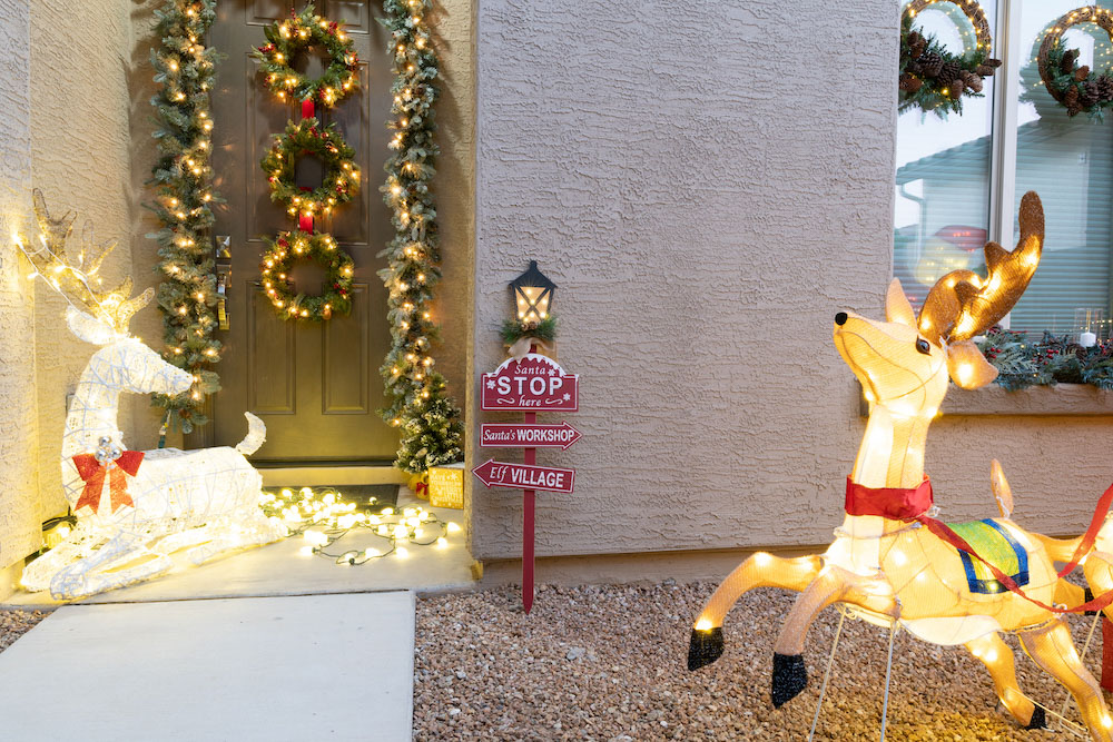 A front porch is styled with a holiday sign, LED deer, garland, wreaths, a small tree, lights and LED Santa display with reindeer  