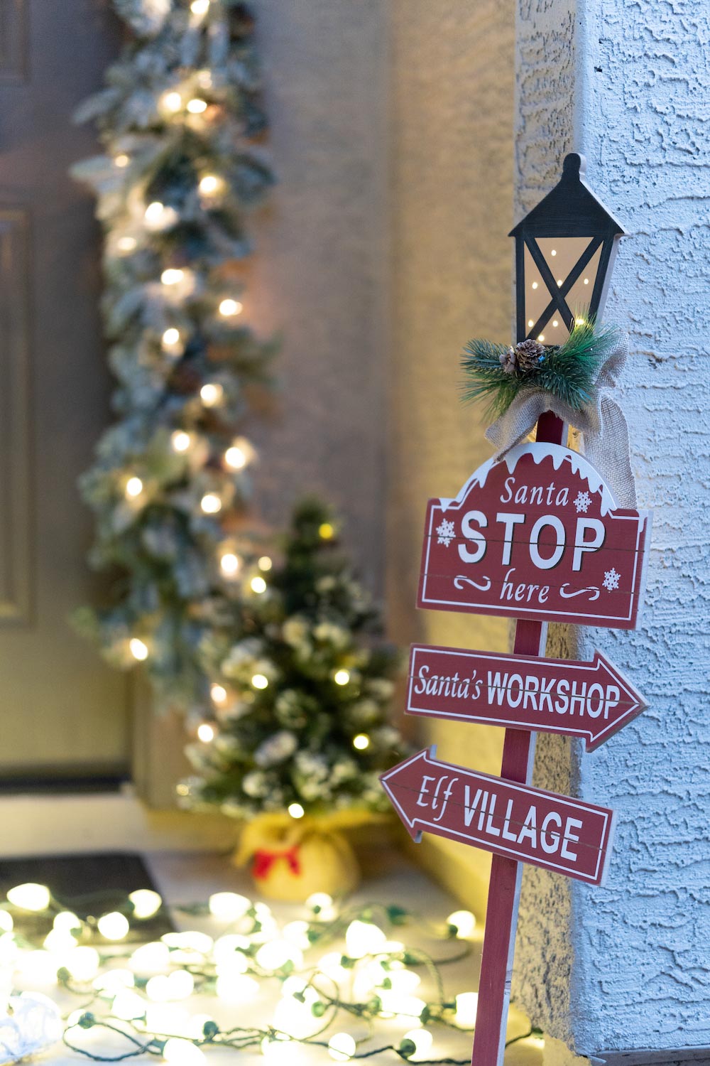 A front porch is styled with a holiday sign, garland, a small tree and lights lit at night