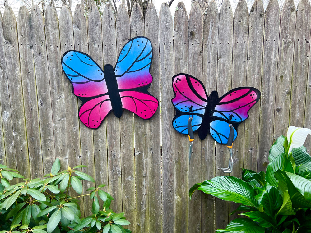 Painted butterflies on a wooden fence, and plants on either side. 