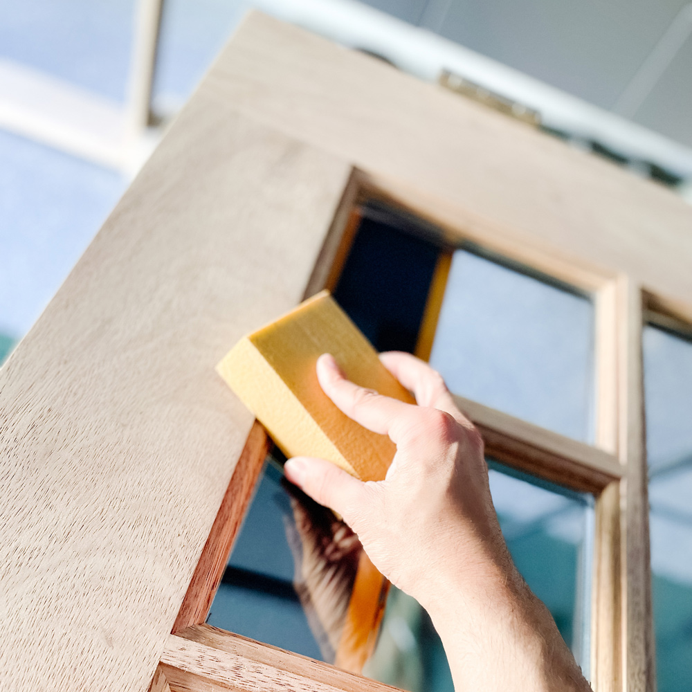 A hand sanding an unfinished Dutch door panel with windows.