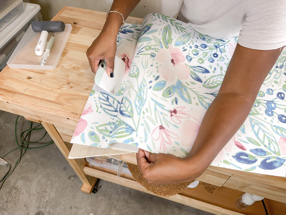 A woman using a scraper tool to flatten floral wallpaper onto a piece of plywood A woman using a scraper tool to flatten floral wallpaper onto a piece of plywood
