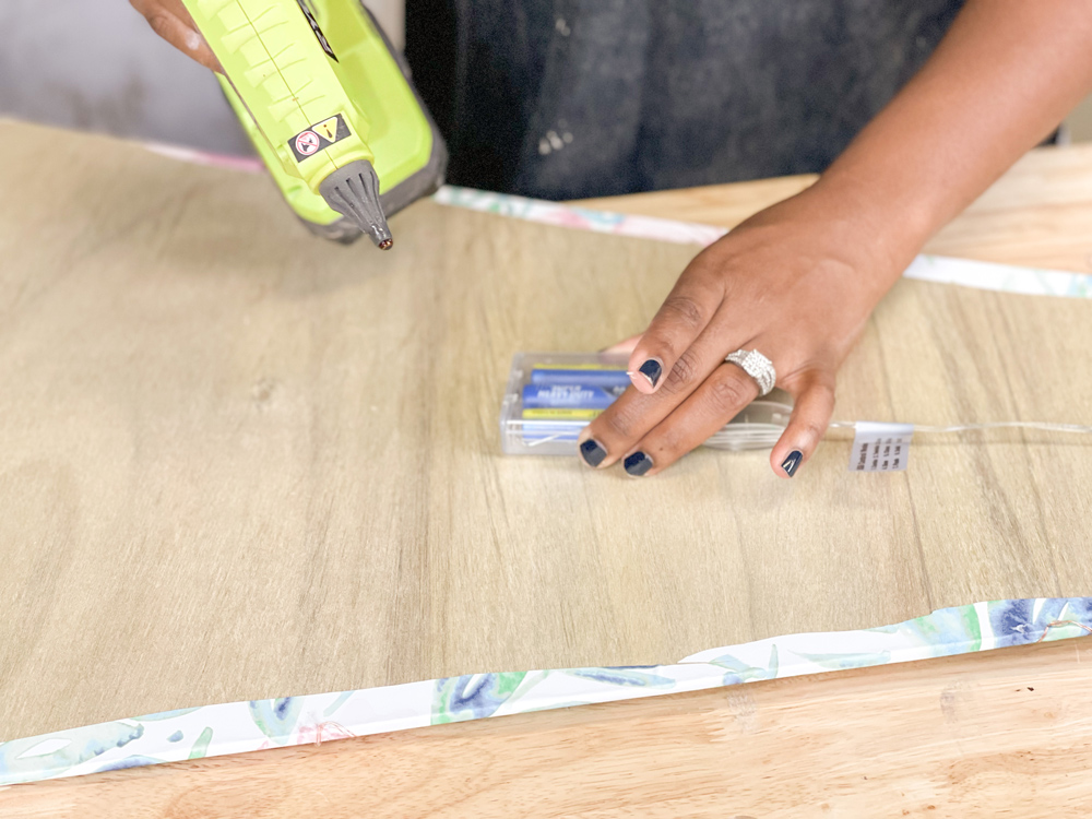 A woman holding a battery pack onto the back of a plywood tree with a green hot glue gun in other hand A woman holding a battery pack onto the back of a plywood tree with a green hot glue gun in other hand