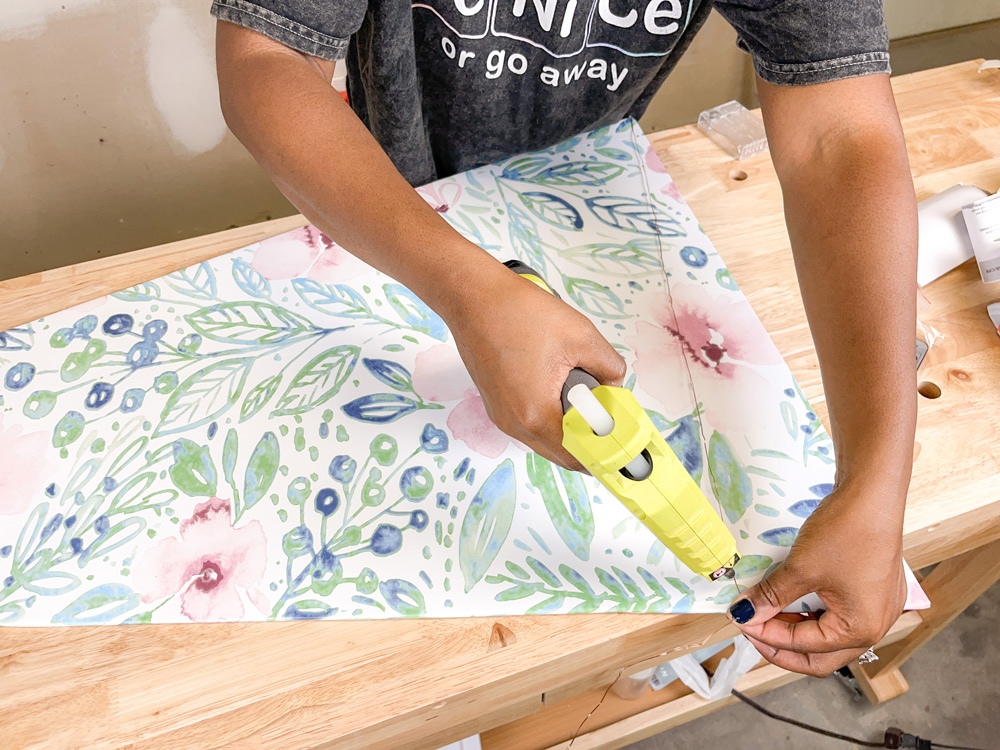 A woman standing over a table and using a green hot glue gun to glue wire onto a wallpapered surface A woman standing over a table and using a green hot glue gun to glue wire onto a wallpapered surface