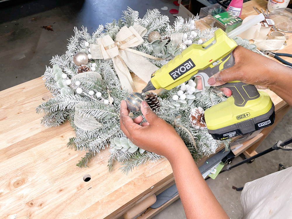 Hands using a hot glue gun to apply glue to a small, grey Christmas bulb.