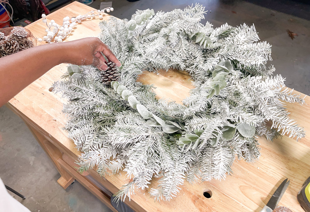 A hand attaching a pinecone to a snowy wreath.