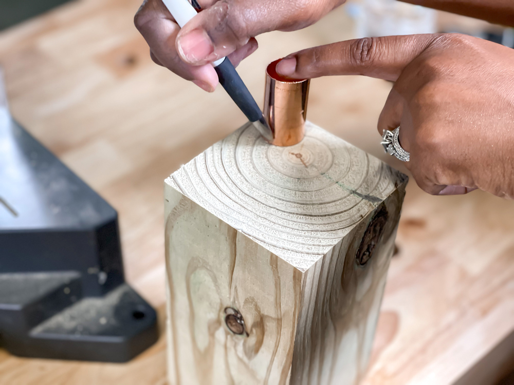  A woman’s hand holding a copper piece on a block of wood with finger, and another hand using a pen to trace the piece