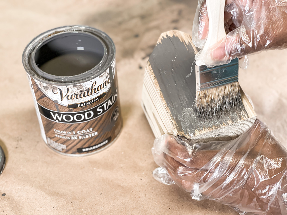 A small can of stain and hands with clear gloves using a paintbrush to stain a piece of wood