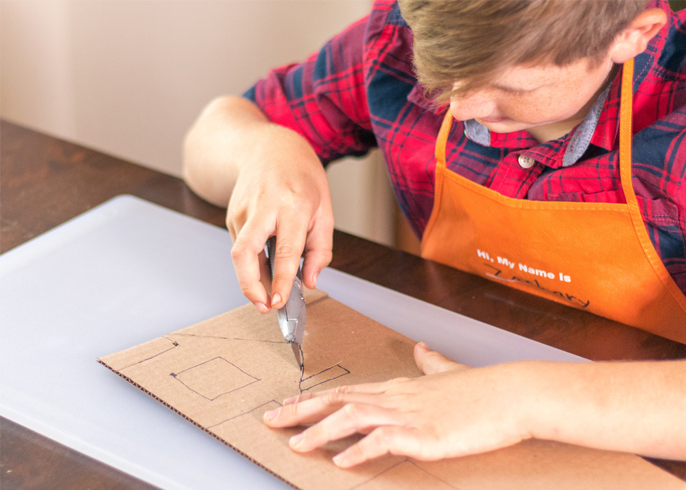 Boy cutting out cardboard pieces with a box cutter.