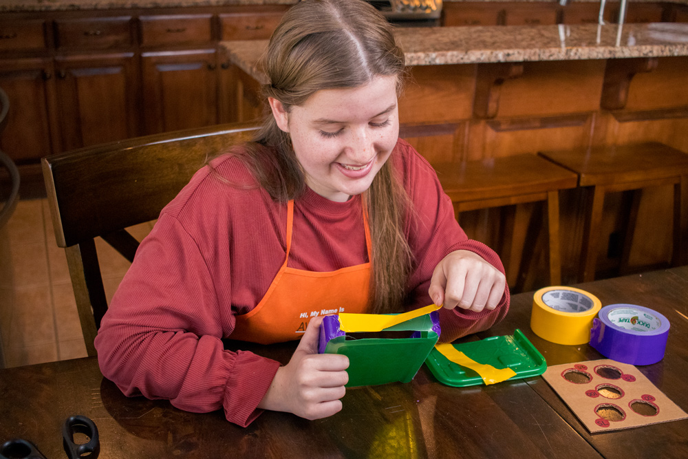 A girl applying colorful duct tape to a cut out green plastic container.
