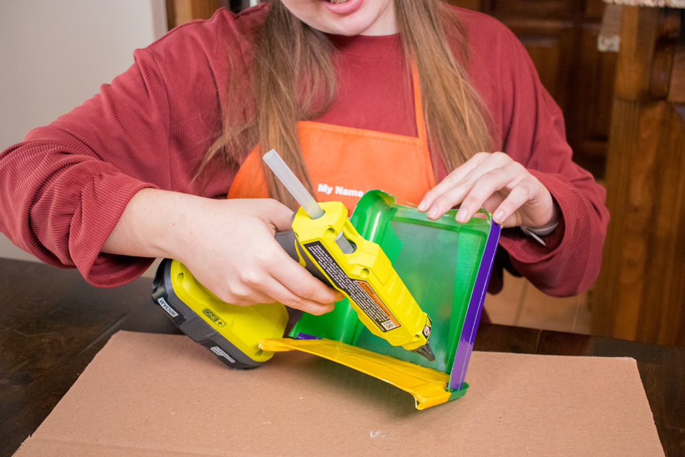 A girl using a hot glue gun to glue a cut out green plastic container.