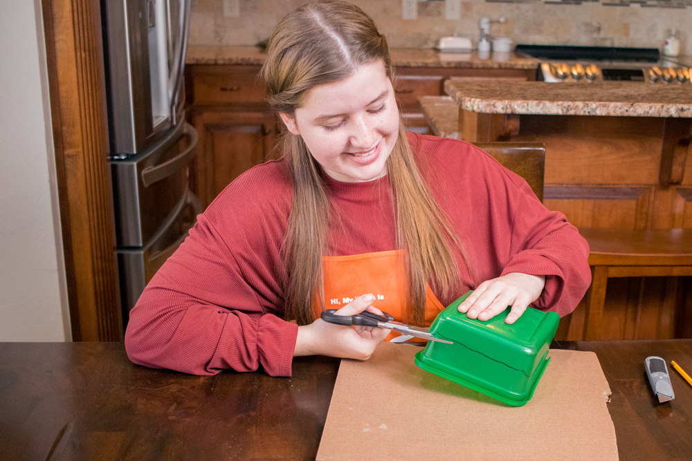 A girl cutting a green plastic container with black scissors.