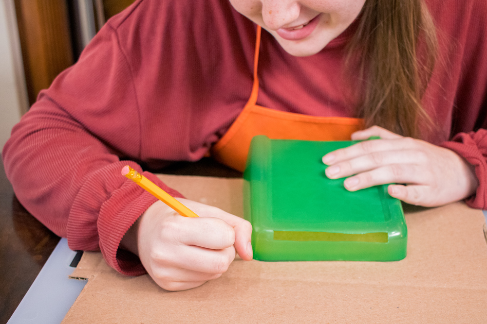A girl using a cut green plastic container to trace on cardboard with a pencil