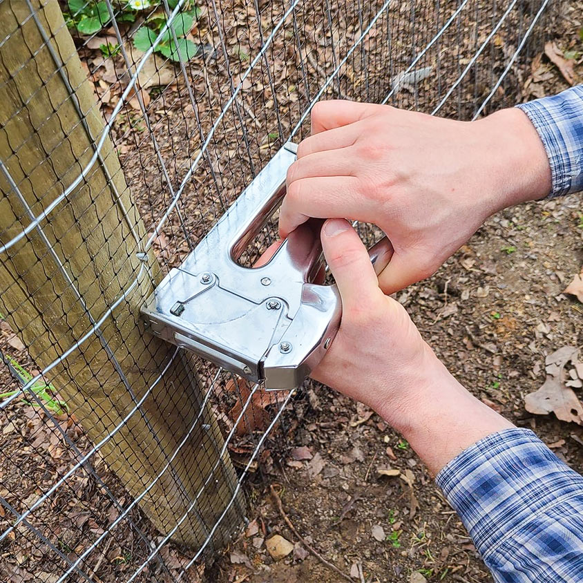 A man stapling wire fencing to a wooden post.