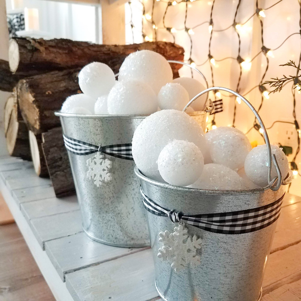 Faux snowballs placed in a silver pale with logs and Christmas lights in the background
