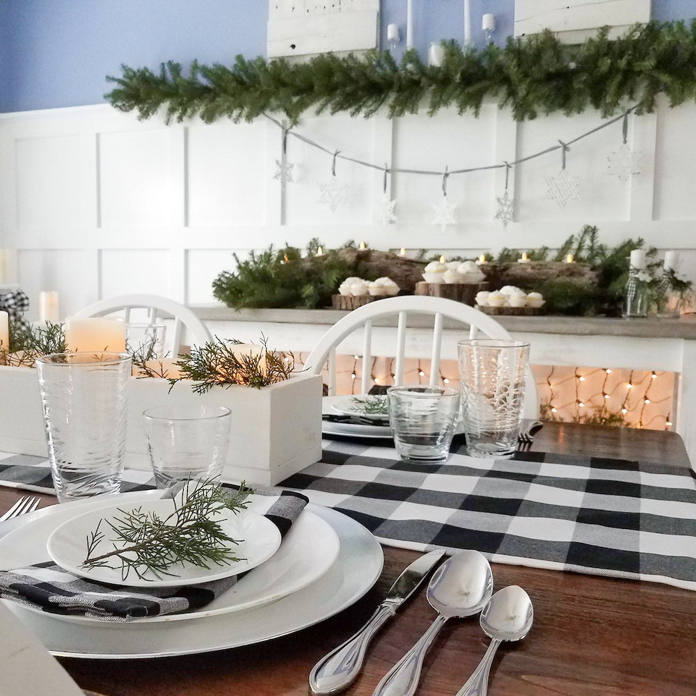Angled image of a dinner table with a black and white table runner and dinner ware with greenery decor in the background