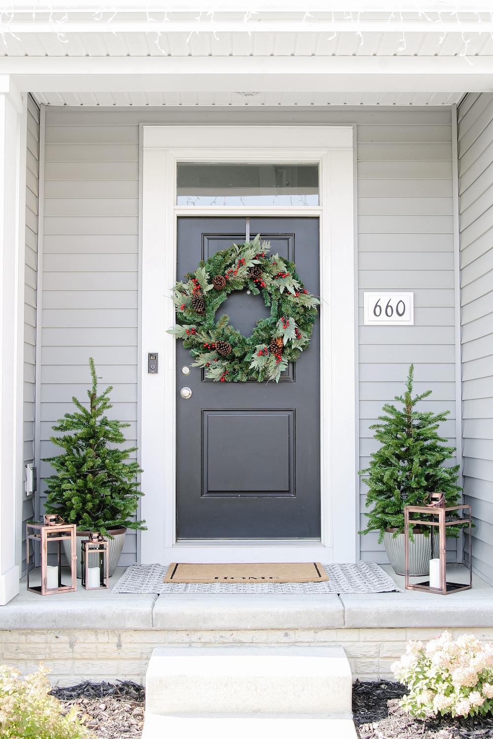 A front porch styled for the holidays with door wreath, porch trees and lanterns